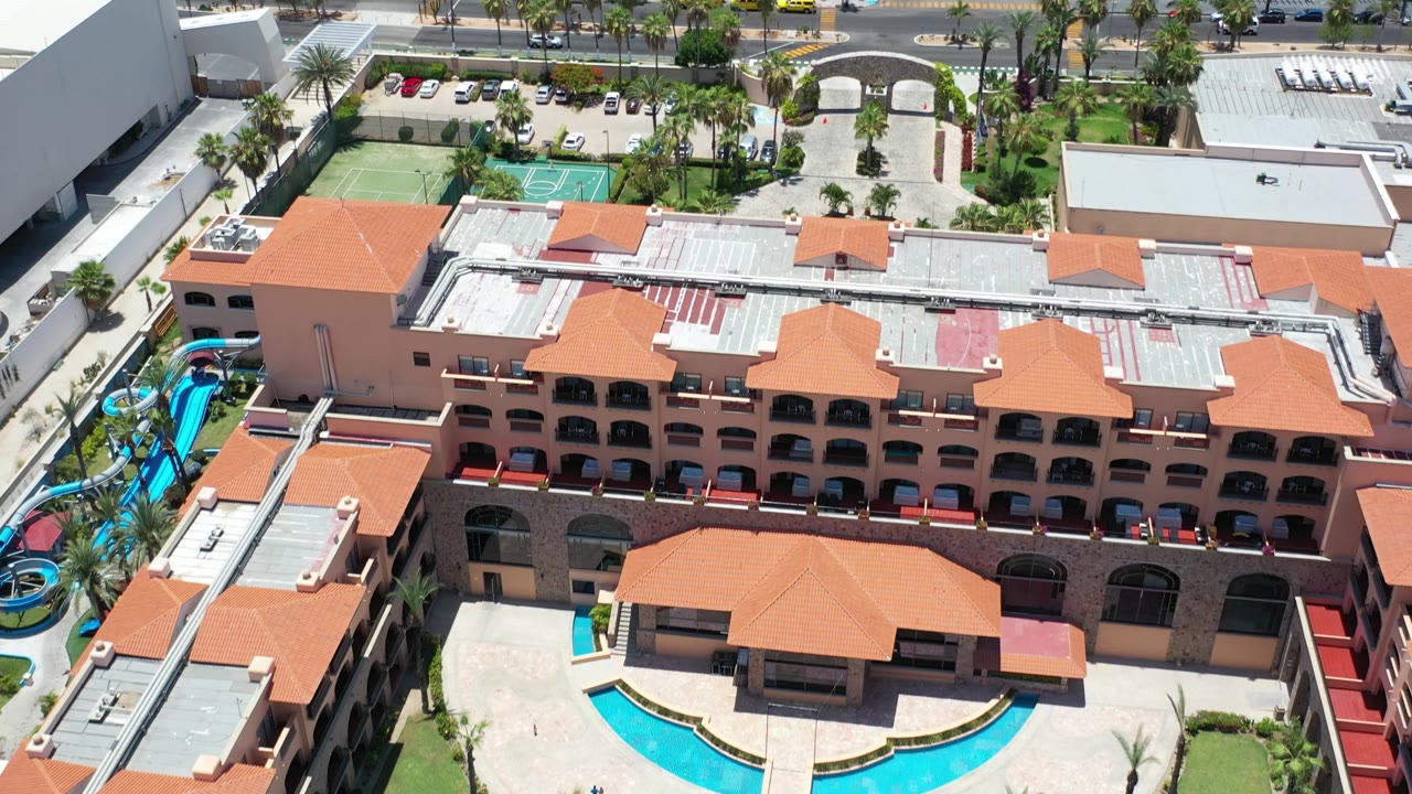 High aerial view in Los Cabos, Mexico of a resort building, pool courtyard, tennis courts, palms, parked cars, and a road beyond the property, with no visible people.