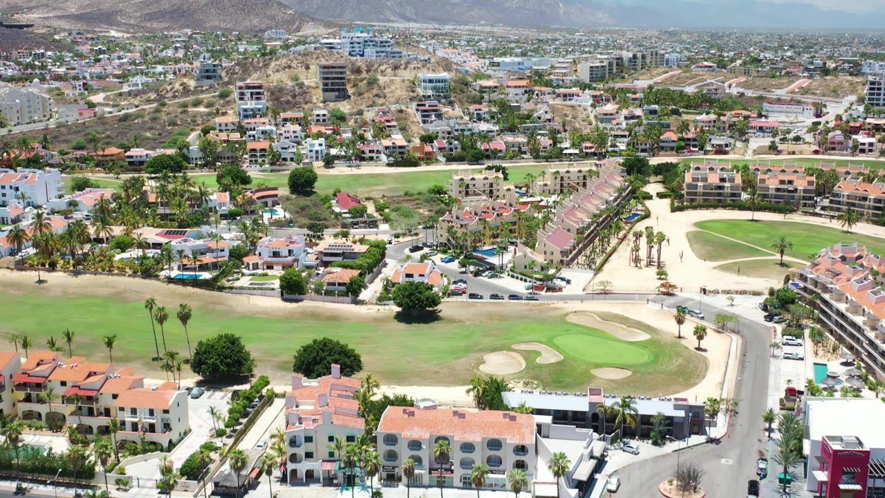 Aerial view over Los Cabos, Mexico, with a curved road beside golf greens, terracotta-roof homes in the foreground, denser hillside buildings in the midground, mountains in back, and no visible people.
