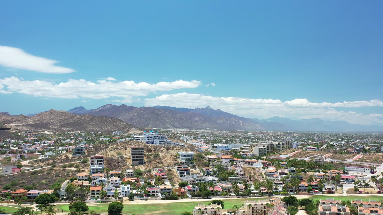 Opening frame of Los Cabos, Mexico, from a high wide angle, with hillside homes, taller modern buildings, distant mountains, white clouds, and no visible people.