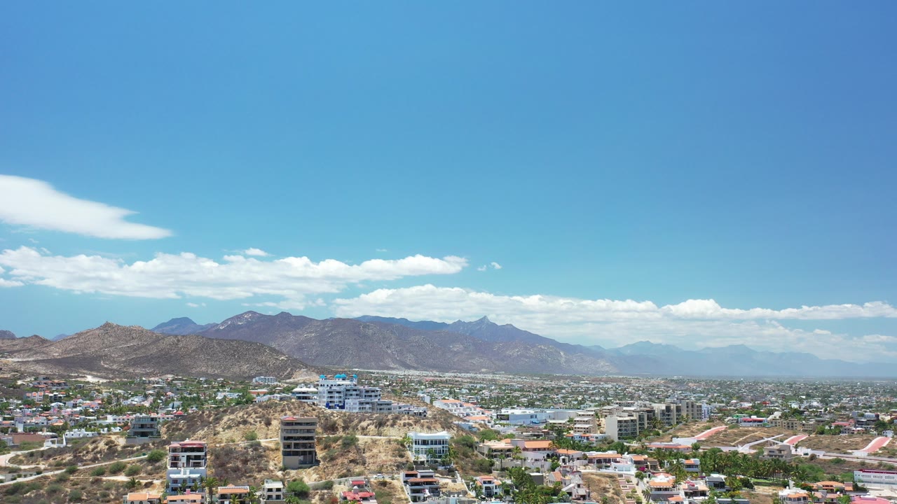 Opening frame of Los Cabos, Mexico from above, with hillside homes in the foreground, a developed valley beyond, distant brown mountains, bright clouds, and no visible people.
