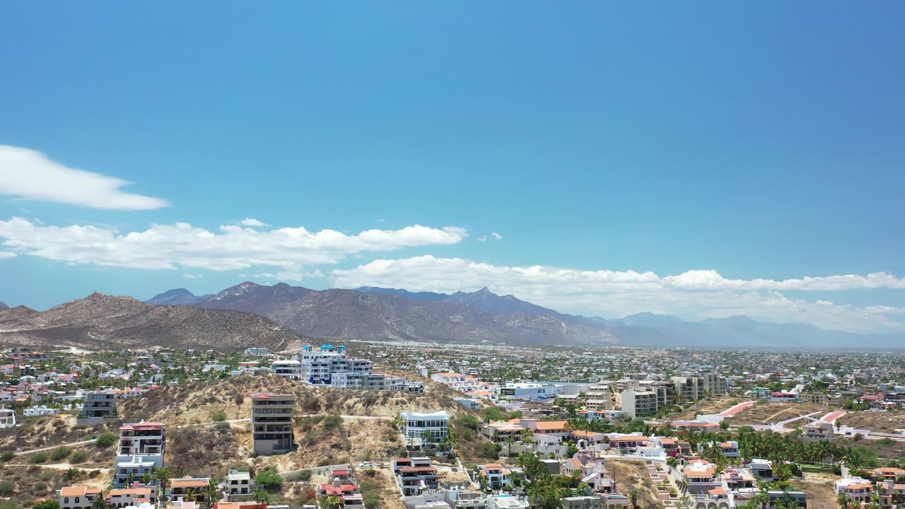 Wide opening aerial view of Los Cabos, Mexico, showing hillside homes, modern buildings, distant mountains, scattered clouds, and no visible people.