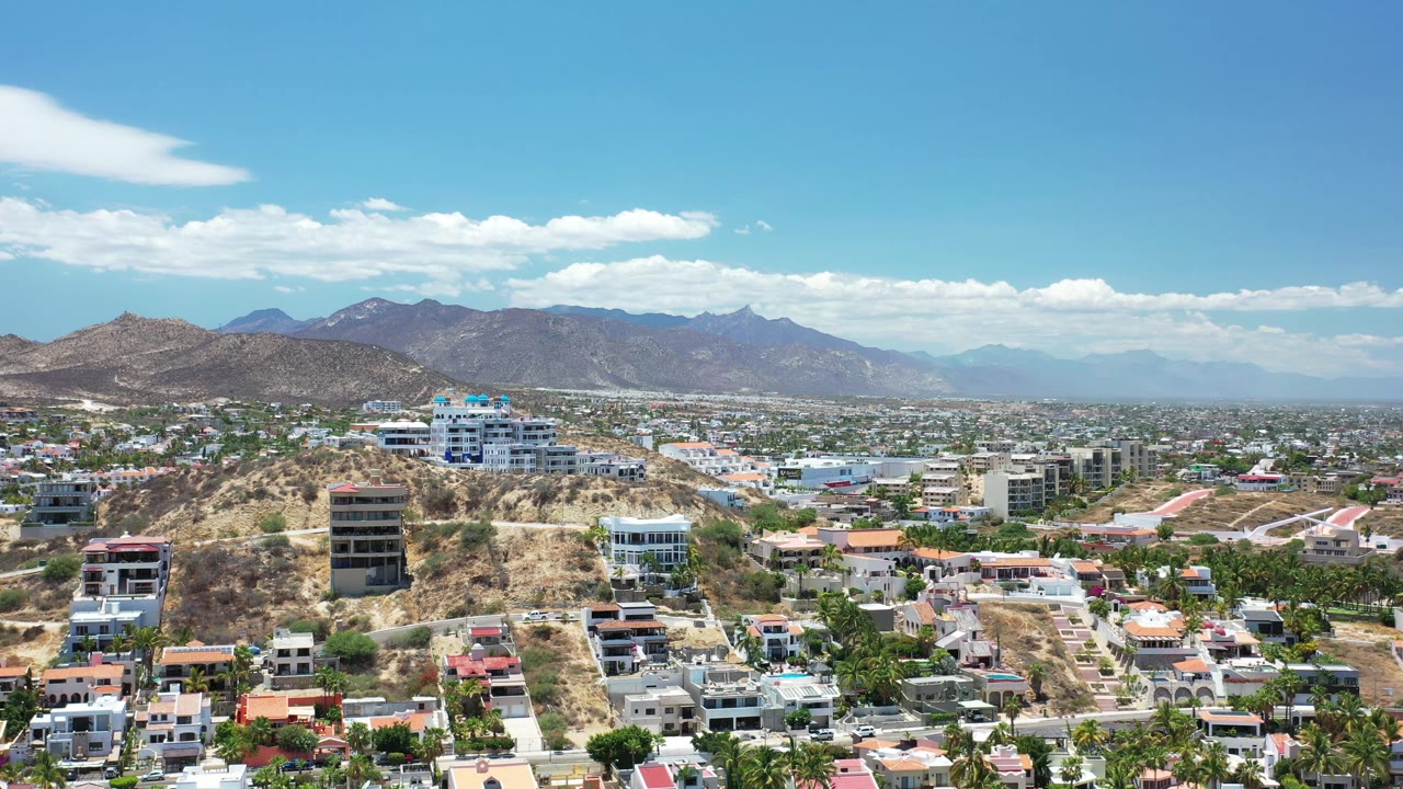 Los Cabos aerial frame with a dry hillside in front, modern homes and streets below, a broad city grid beyond, mountains on the horizon, and no visible people.