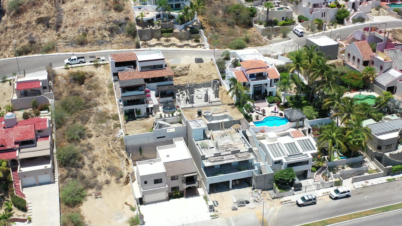 Elevated aerial view over modern homes in Los Cabos, Mexico, with dry hillside lots, palm trees, paved streets, parked vehicles, and no visible people.