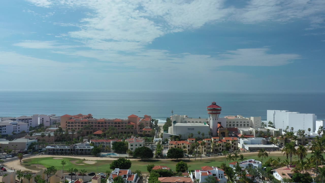 High aerial view over resort buildings and palms in Los Cabos, Mexico, with a wide blue ocean horizon and no visible people.