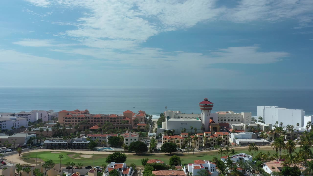 Aerial frame of a Los Cabos resort district with red-roofed buildings, white hotel-like structures, palms, ocean, and no visible people.