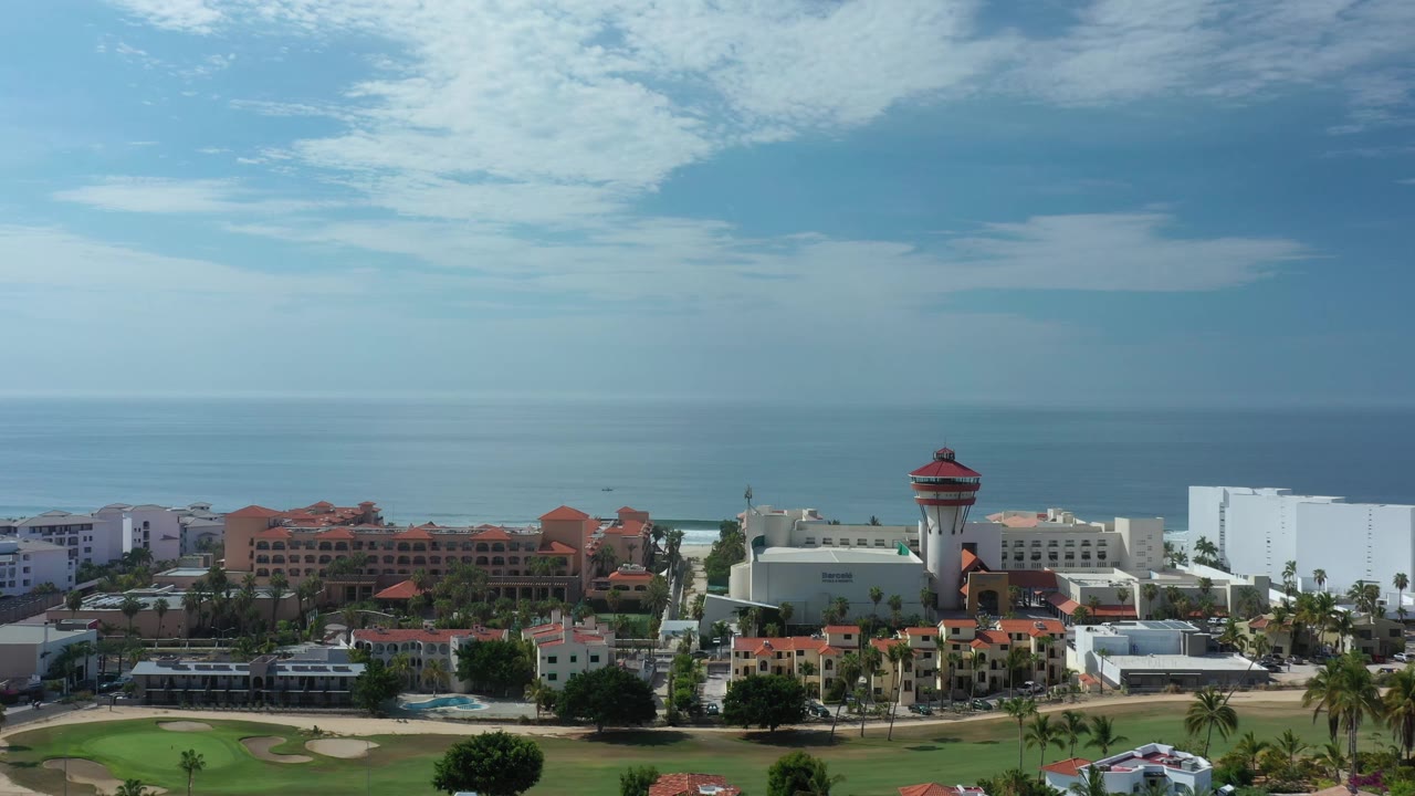 Early aerial frame over Los Cabos, Mexico, with the red-capped tower near center right, white buildings, red-roofed condos, ocean, and no visible people.