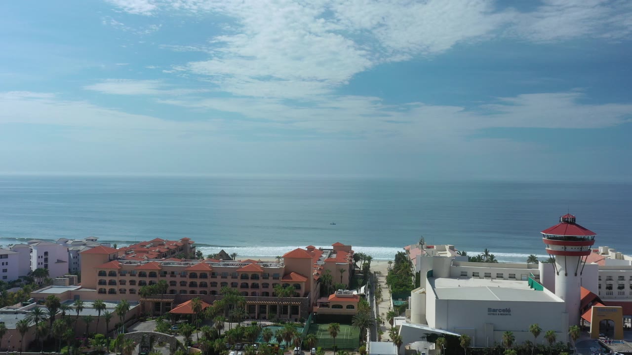 Los Cabos coastal view with red-roofed hotel buildings, a white tower, beach surf, open ocean, scattered clouds, and no visible people at the start of the clip.