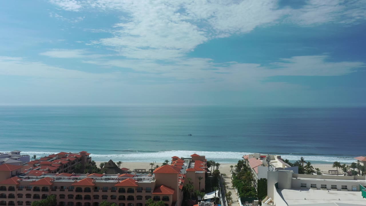 Los Cabos coastline seen from above with calm blue water, a thin breaking-wave line, red roof clusters in the foreground, and no visible people.