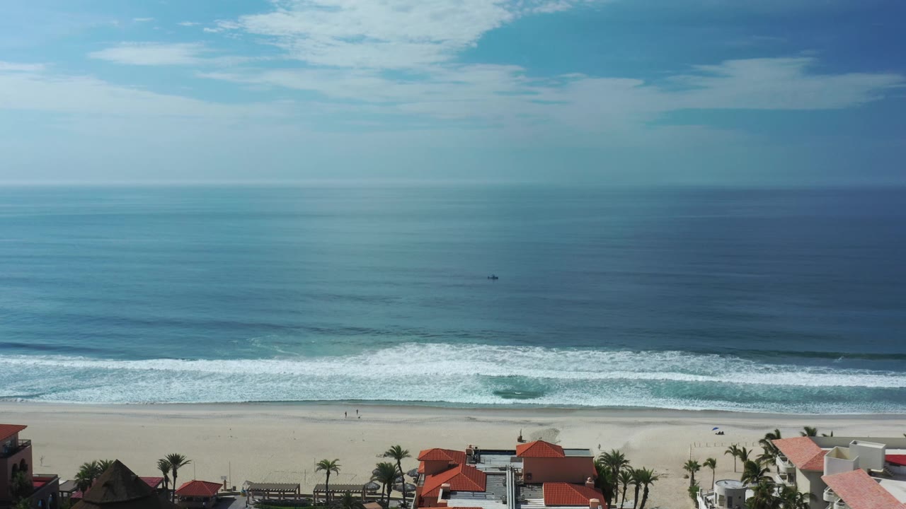 Los Cabos beach view one second in, with the surf line slightly farther across the frame, blue water filling most of the image, and small beachgoers near the waterline.