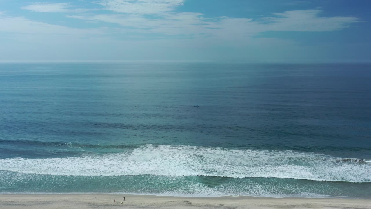 Wide aerial view of dark ocean waves meeting a sandy beach under blue sky.