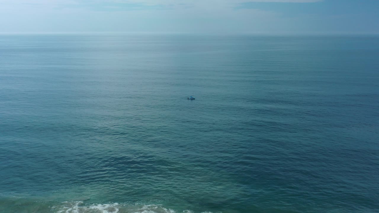 Wide aerial view over blue-green ocean near Los Cabos, Mexico, with a tiny dark person or object offshore and faint surf near the lower edge.