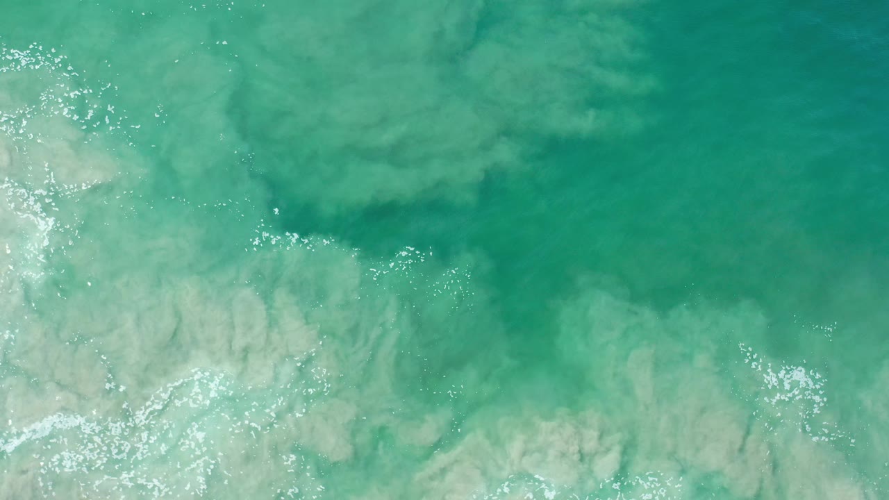 Green-tinted seawater over the pale seabed in Los Cabos, Mexico, with bright surface glints near the left edge and no visible people.