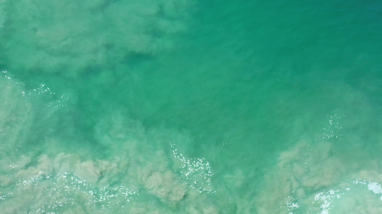 Overhead view of turquoise ocean water in Los Cabos with tan sand plumes near the lower edge, scattered white foam, and no visible people.