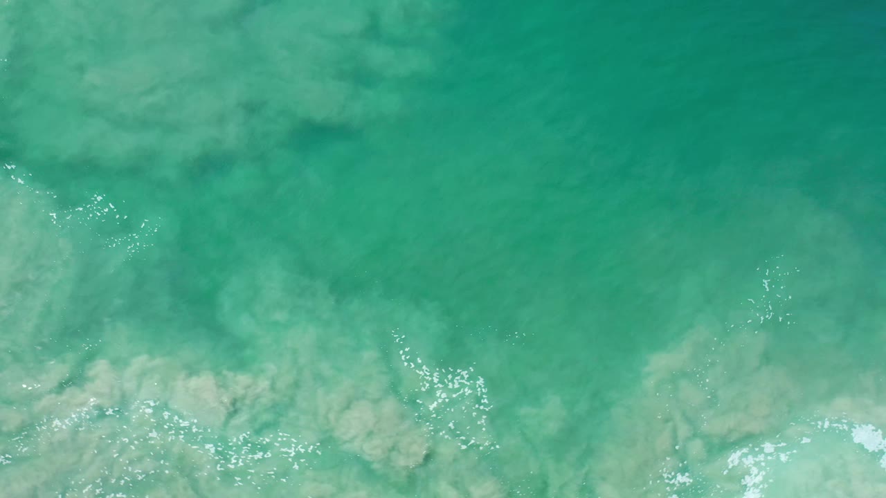 Overhead Los Cabos ocean frame with cloudy tan silt spreading through blue-green water, bright reflection specks, and no visible people.