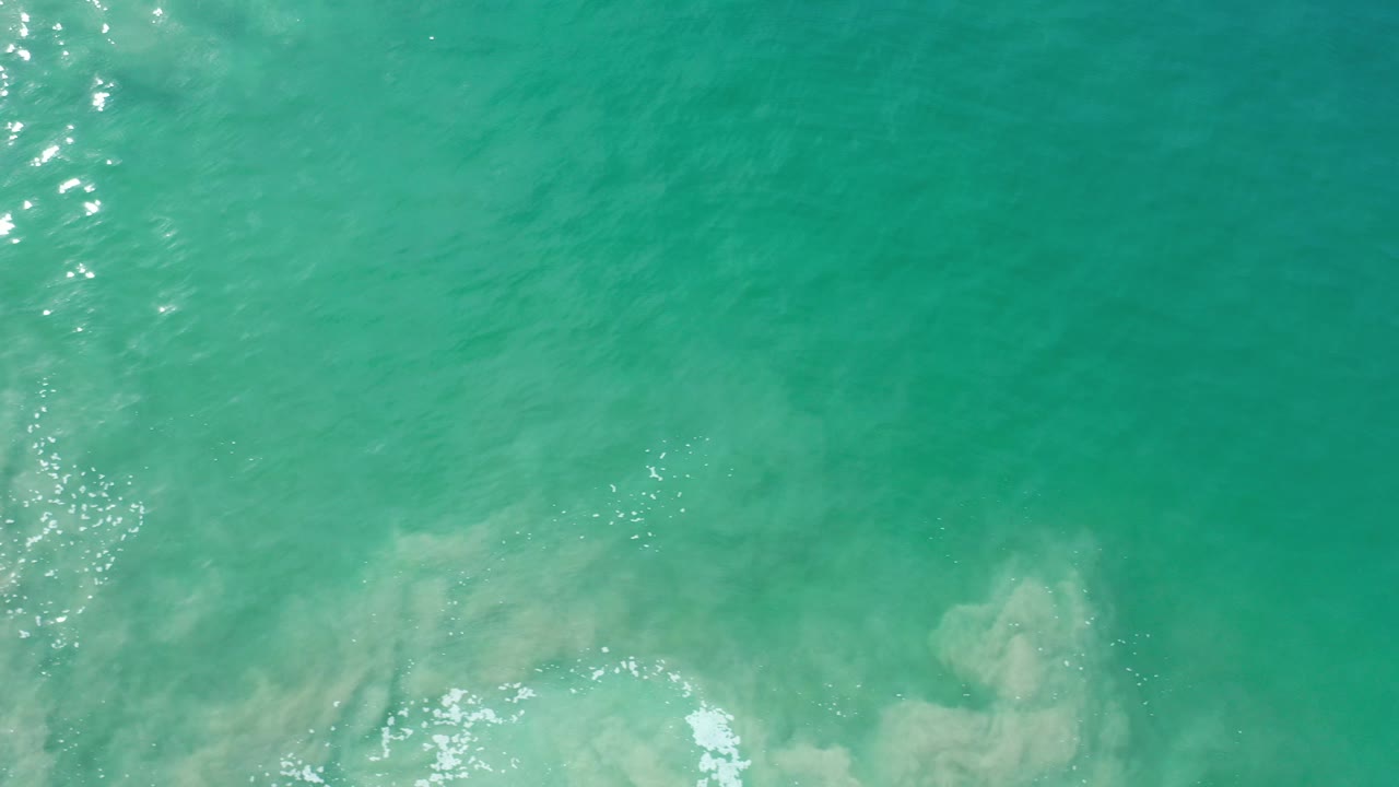 Overhead view of turquoise water in Los Cabos, Mexico, with pale underwater sand or rock forms, small white foam at the bottom, bright sun glints at left, and no visible people.