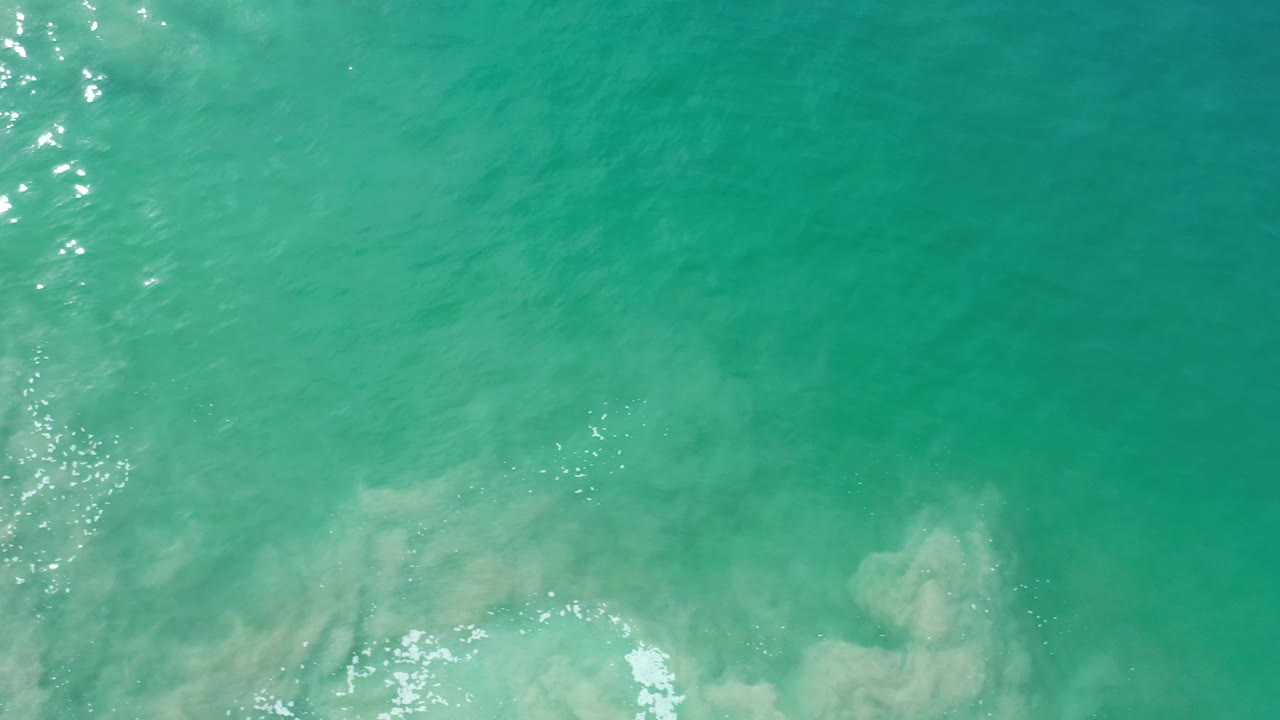 Overhead Los Cabos water frame with turquoise surface, pale submerged forms near the lower half, white foam along the bottom edge, vertical sun reflections at left, and no visible people.