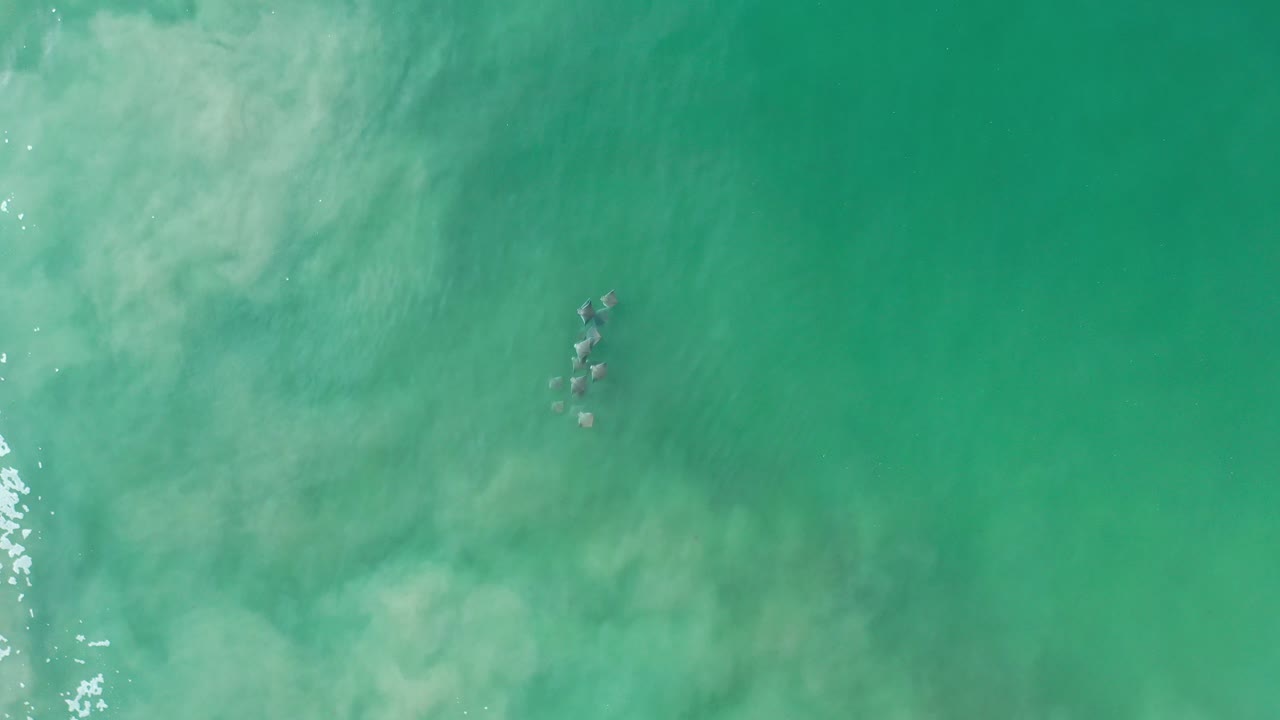 Overhead Los Cabos water view with green-blue tide, a small cluster of submerged rocks near the upper center, pale surf haze, and no visible people.