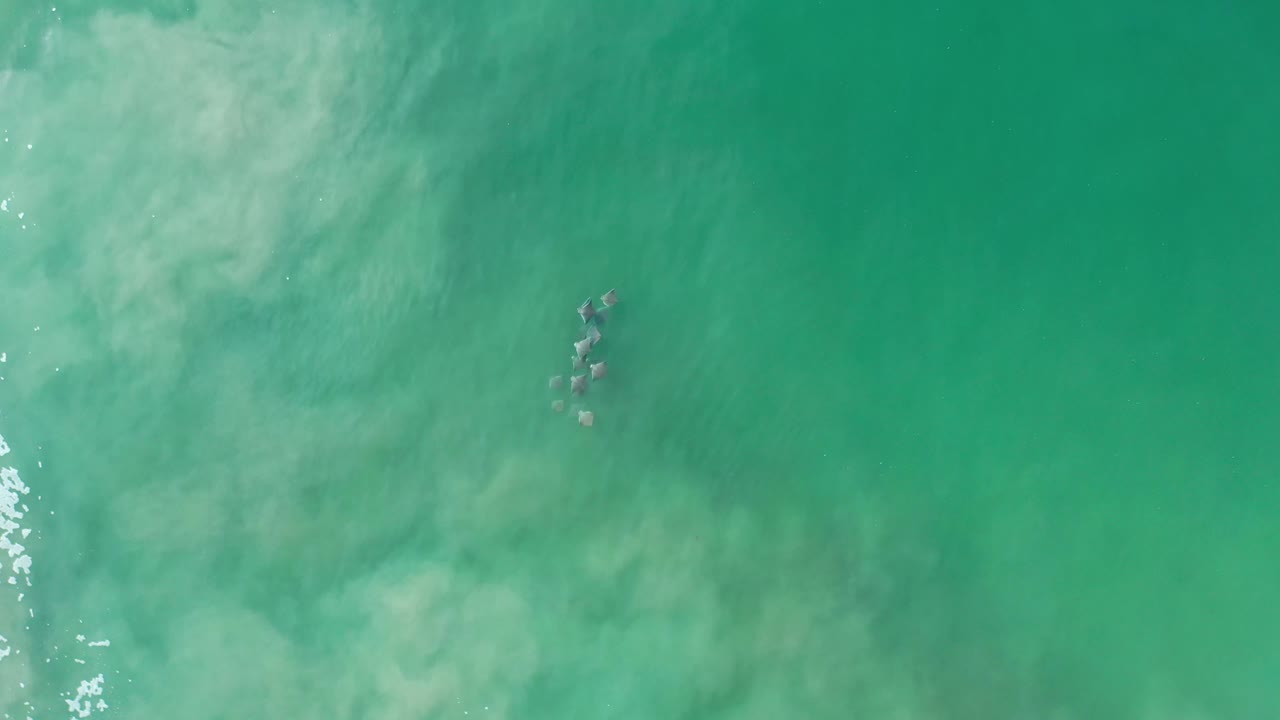 Overhead Los Cabos frame with cloudy pale water spreading across the upper left, dark rocks near center, and no visible people.