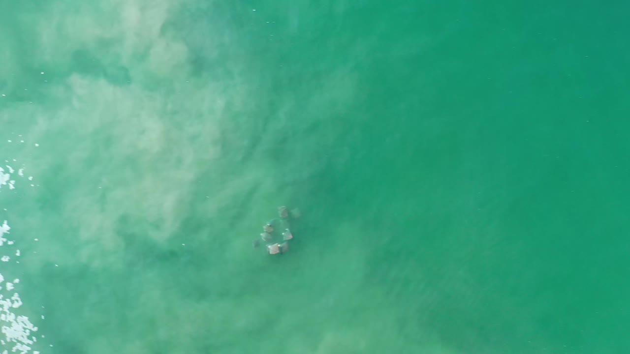 Aerial view of Los Cabos water with a small dark submerged cluster near center and pale sediment drifting through greenish blue water, with no visible people.