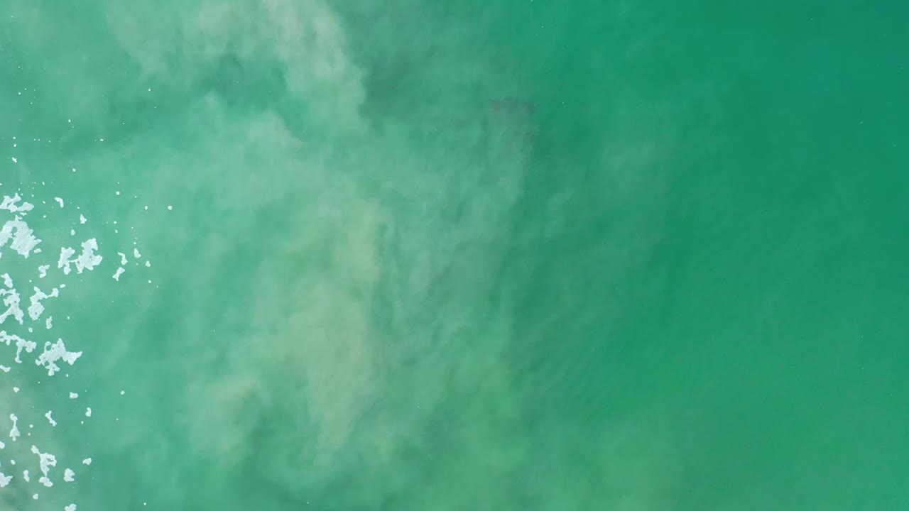 Overhead Los Cabos ocean frame with pale cloudy water patterns and a compact patch of bright ripples just left of center, with no visible people.