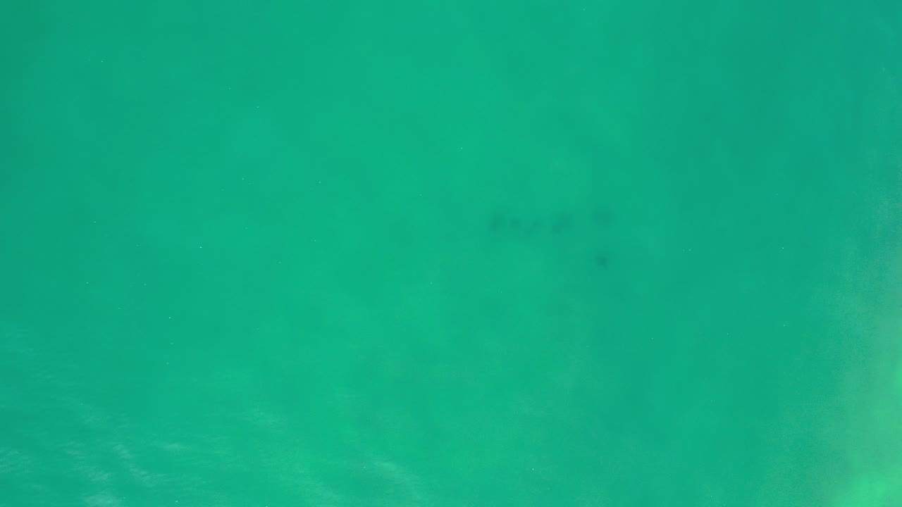 Top-down aerial view of greenish-blue water in Los Cabos, Mexico, with soft ripples, a few darker underwater marks, and no visible people.
