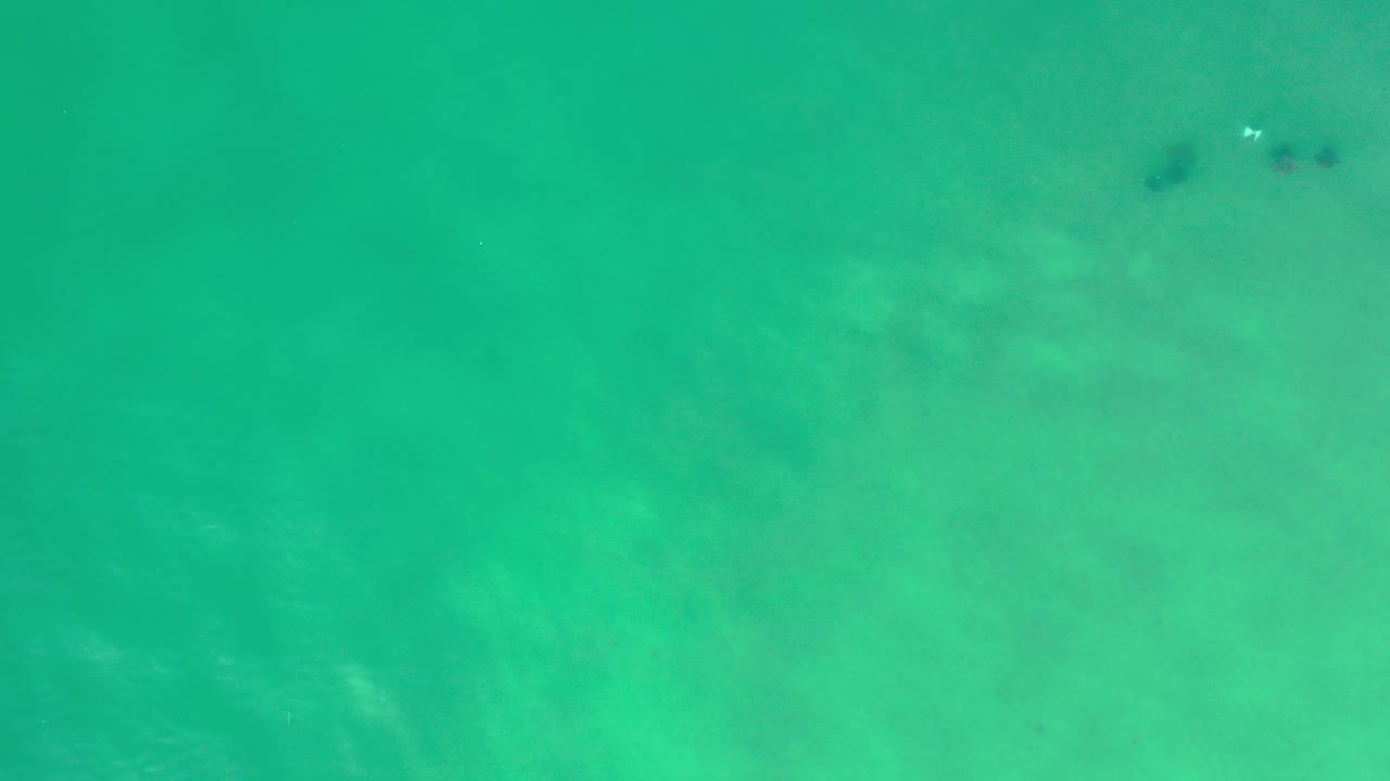 Top-down Los Cabos water frame with green-blue ocean over pale sand, faint wave marks, small dark patches near the top, and no visible people.