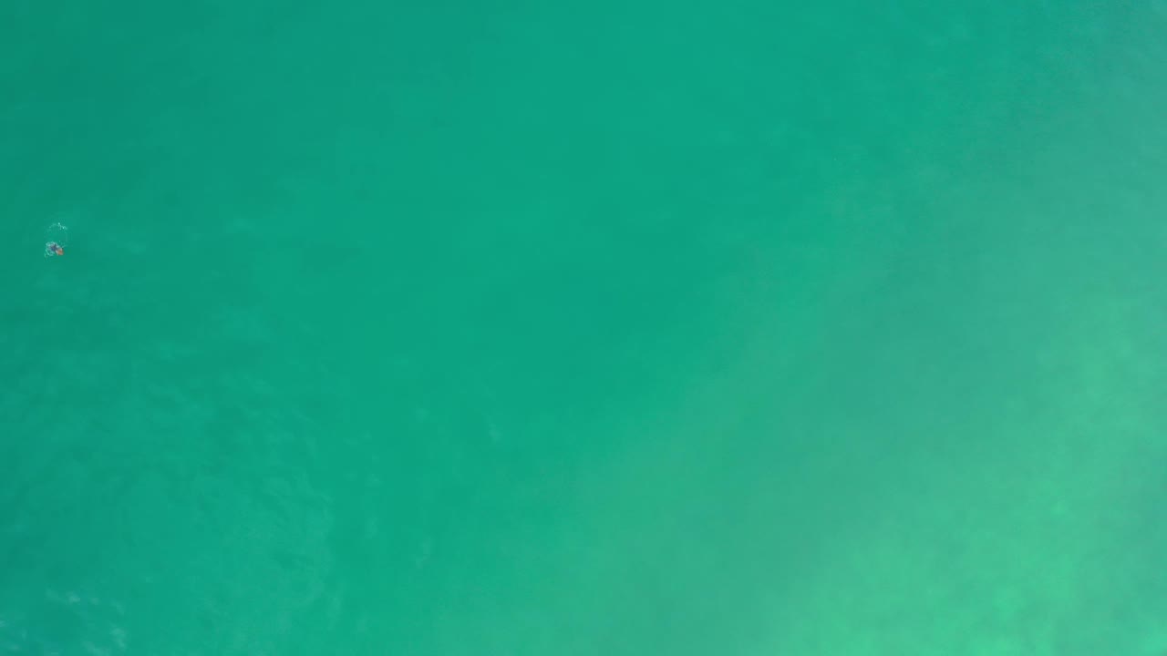 Overhead Los Cabos ocean frame with teal water, soft tonal variation, and a tiny white foam mark near the upper area, with no visible people.