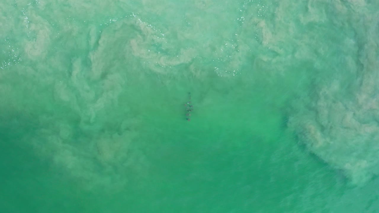 Top-down view of green-blue water in Los Cabos, Mexico, with pale underwater swirls, scattered surface sparkle, a small dark submerged object near the lower center, and no visible people.