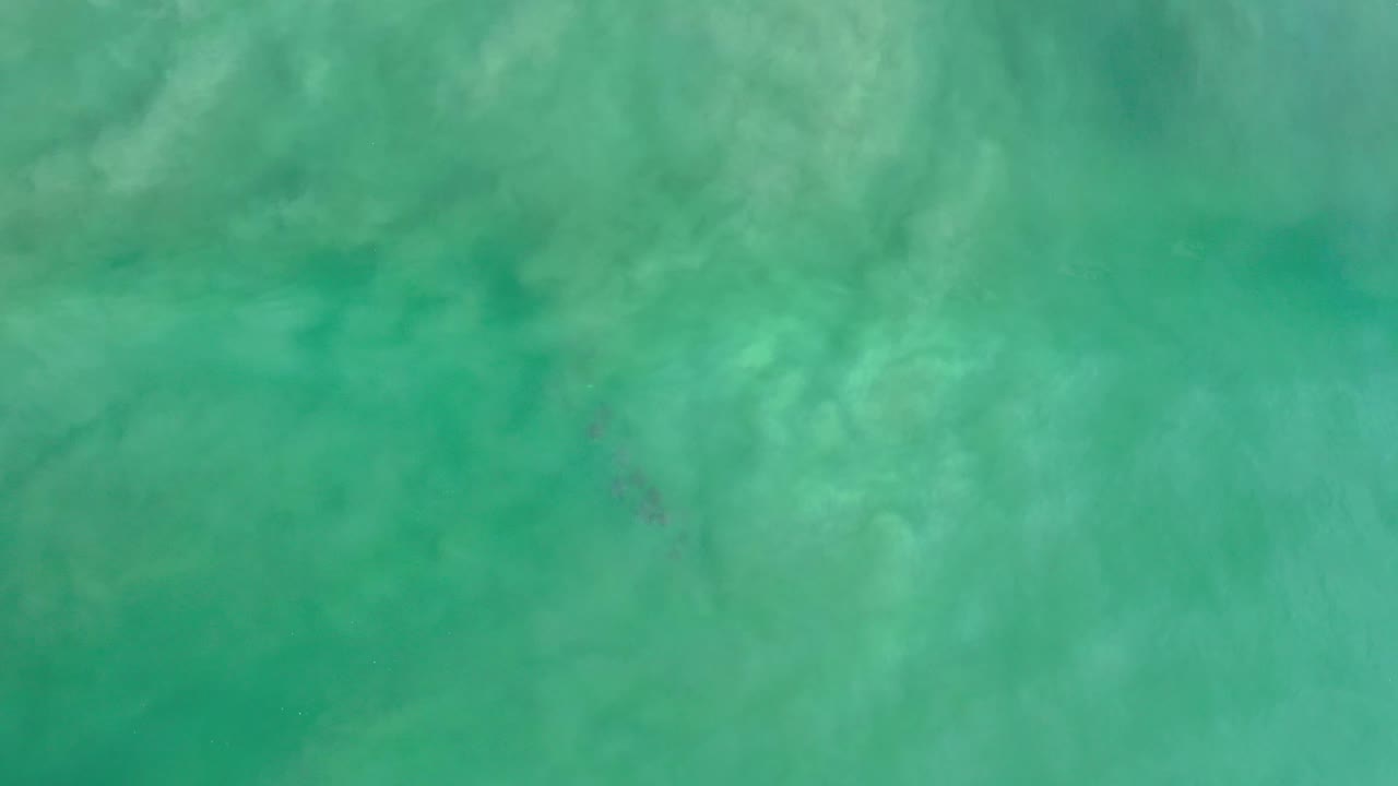 Turquoise Los Cabos ocean fills the frame as cloudy wave wash spreads near a small dark island or reef form, with no visible people.