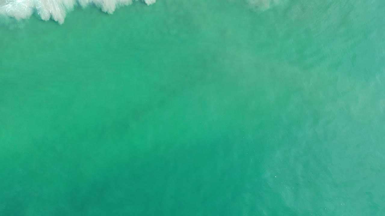 Overhead view of turquoise ocean water in Los Cabos, Mexico, with a narrow line of white foam entering from the top edge and no visible people.