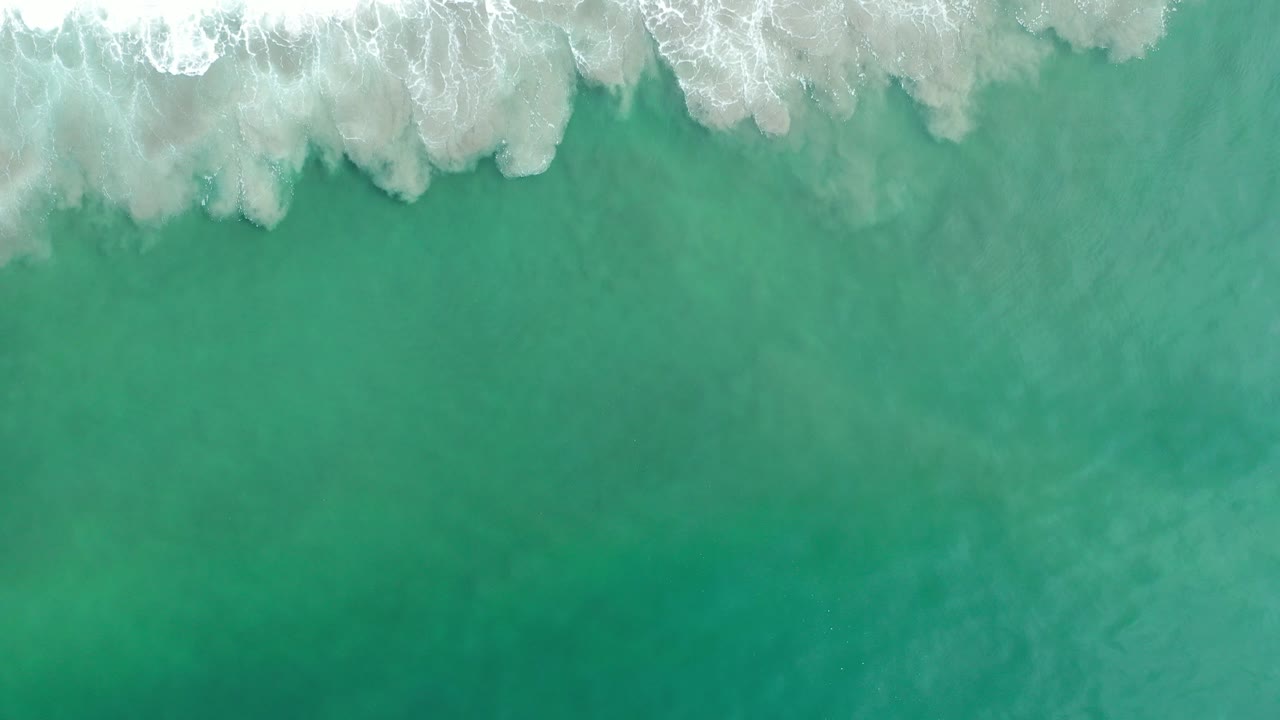 Overhead view of turquoise-green ocean near Los Cabos with a band of white surf entering along the top edge and no visible people.
