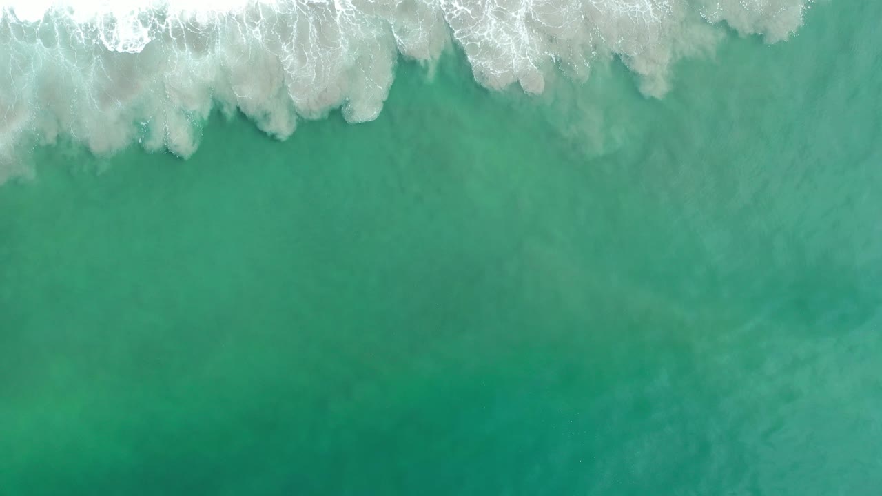 Overhead Los Cabos ocean frame showing white wave foam spreading farther across green water, with no visible people, boats, or shoreline structures.