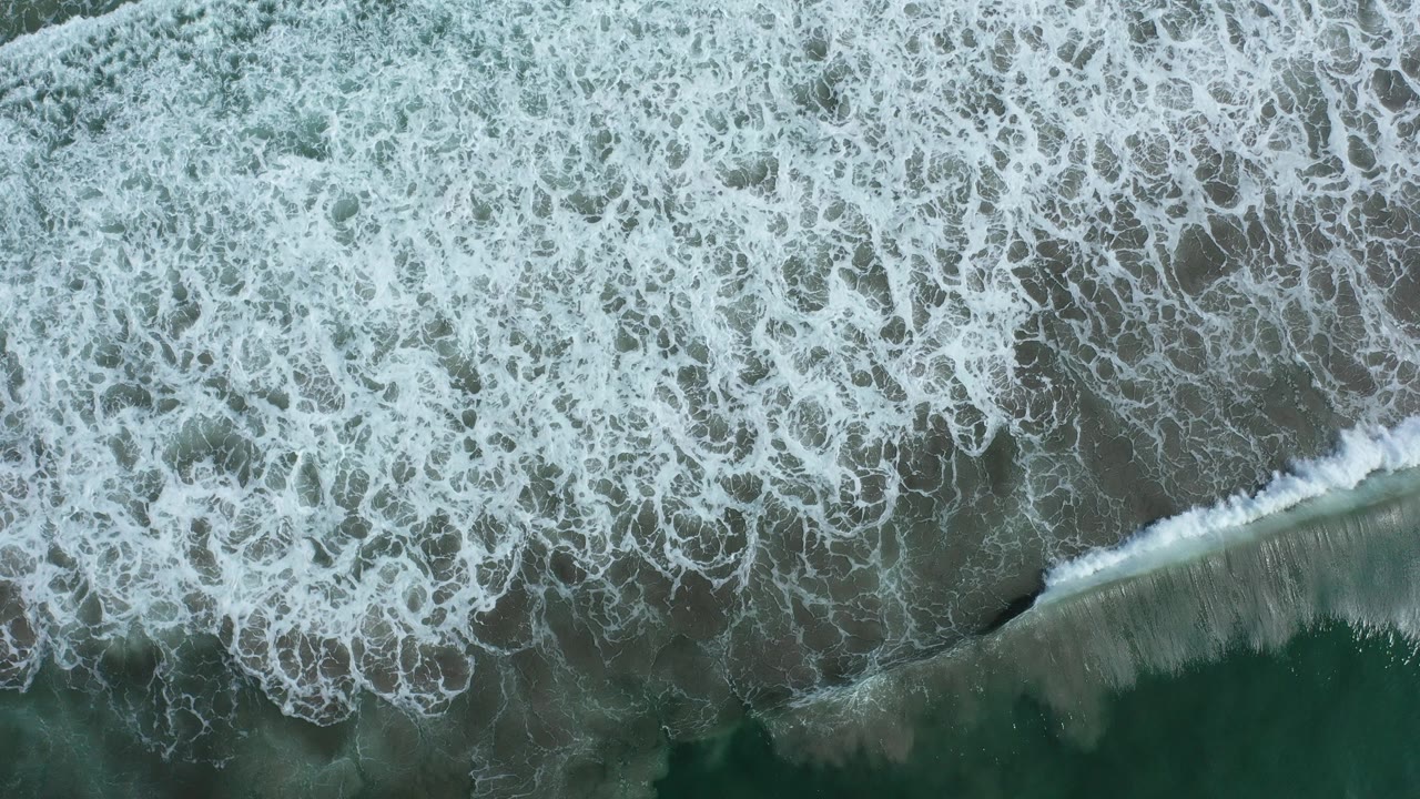 Top-down view of Los Cabos surf where white foam covers most of the frame and a narrow band of wet sand appears at the lower left, with no visible people.