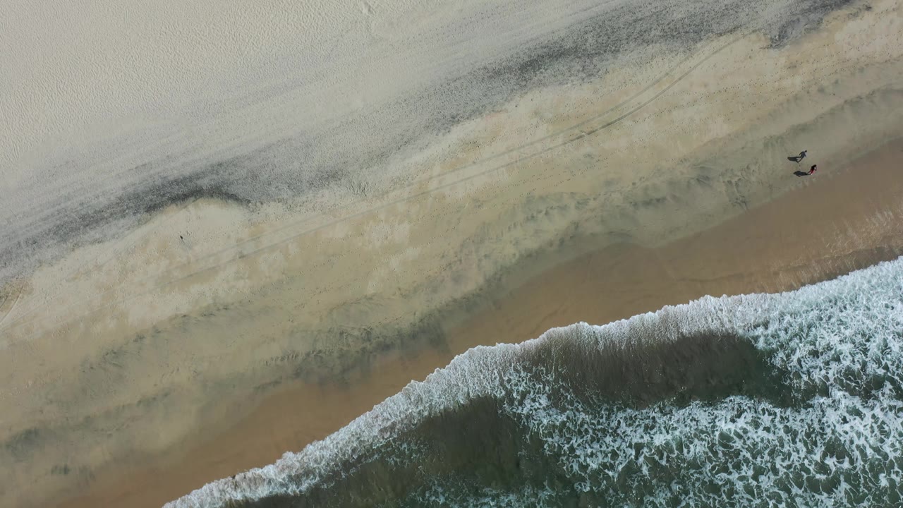 Aerial frame over Los Cabos sand with a curved strip of foam at the bottom edge, darker wet sand in the middle, and open dry beach above.