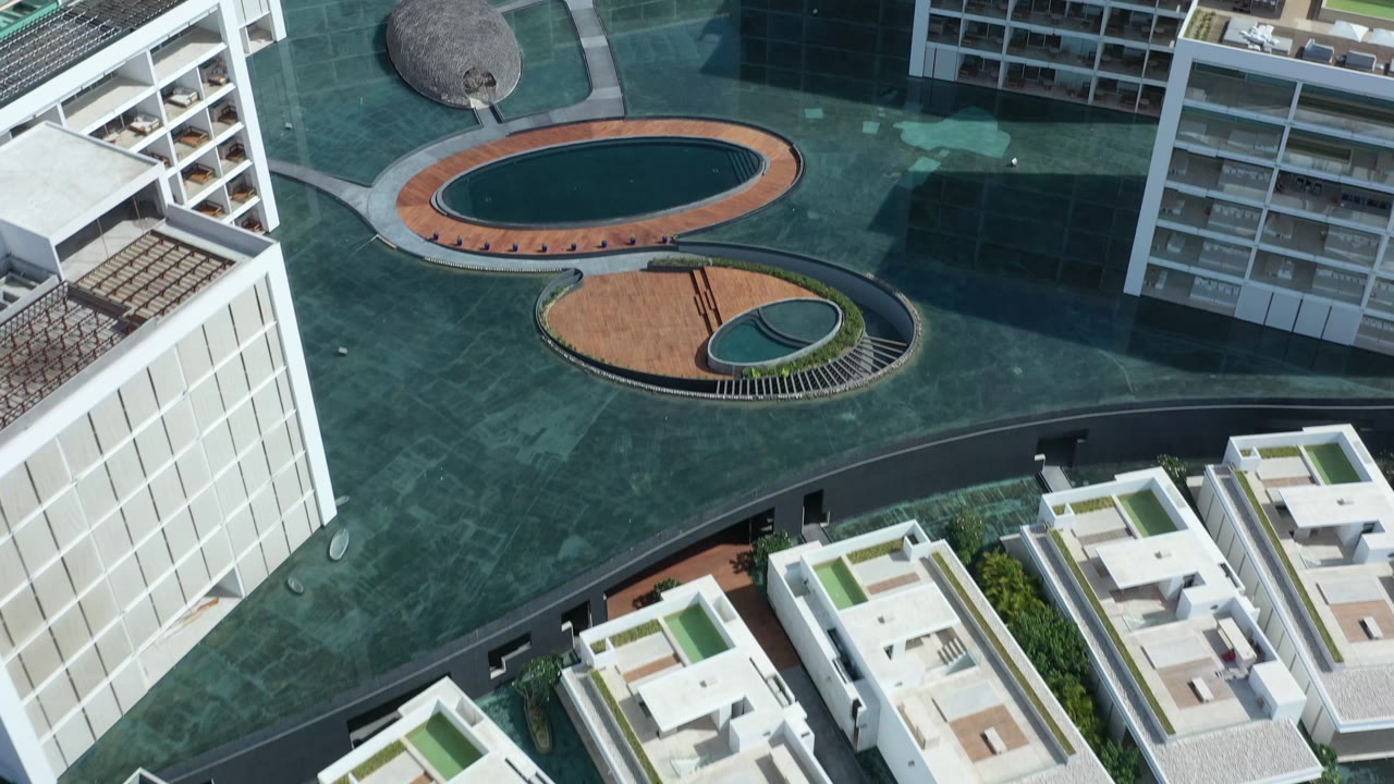 Aerial frame over Los Cabos residential buildings, with white facades, rooftop patio areas, green pavement, dark oval water features, and no visible people.