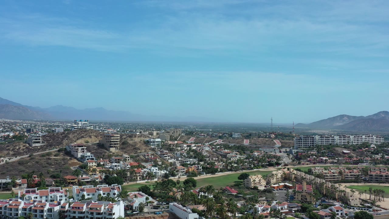 Wide aerial frame over Los Cabos, Mexico, showing neighborhoods, roads, dry hills, distant mountains, hazy blue sky, and no visible people.