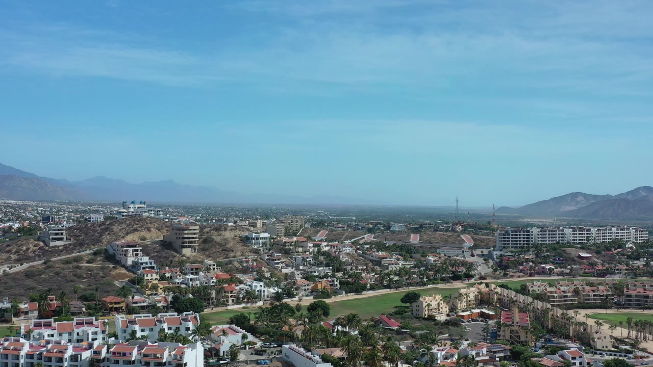 High view over mixed housing, a green field, dry ridges, and distant mountains.