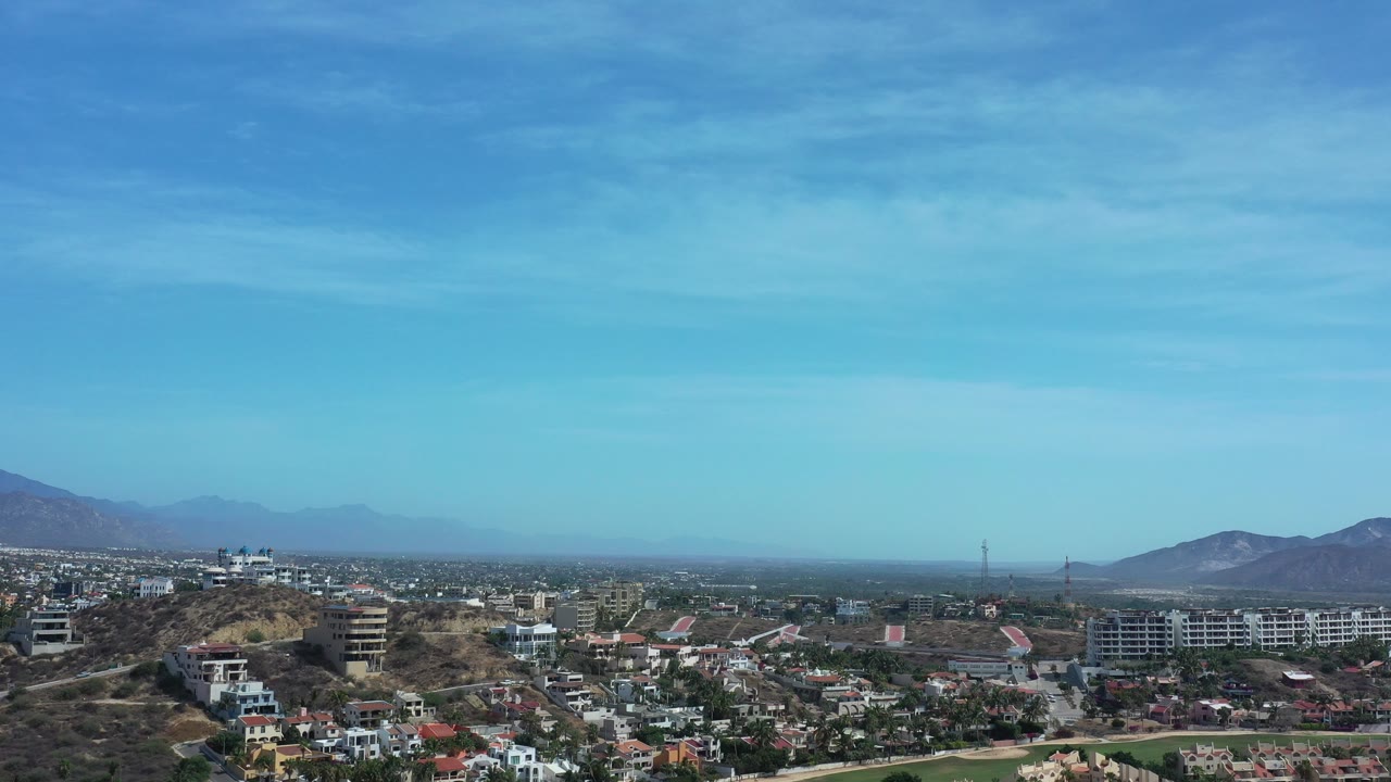 Wide aerial view of hillside homes, green grass, distant mountains, and blue sky.