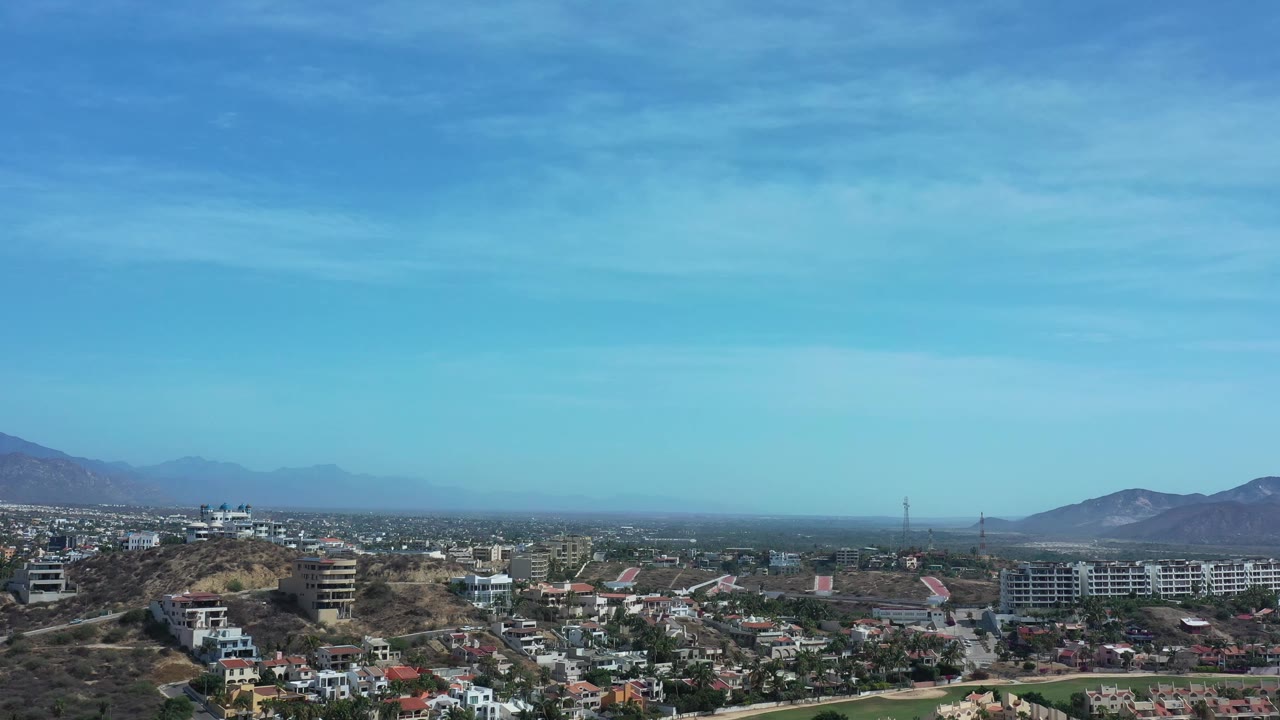 Aerial view in Los Cabos, Mexico, looking across suburban homes, dry ridgelines, distant mountains, and pale blue sky with no visible people.