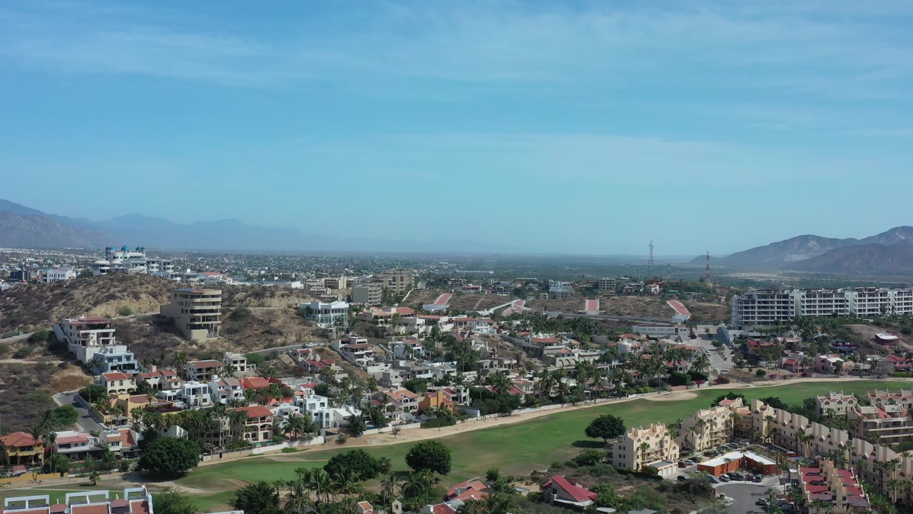 Aerial view over green landscaped ground, homes, hills, and distant mountains.