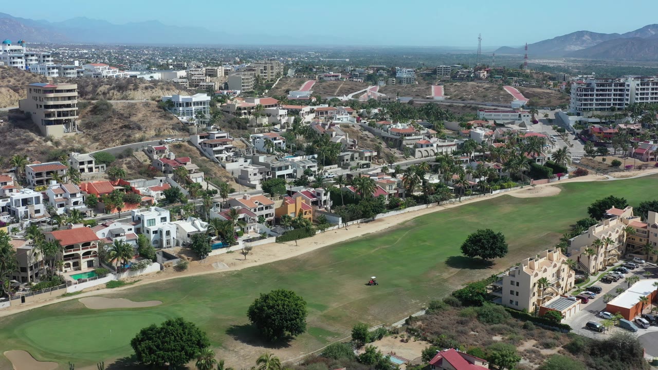 Los Cabos aerial view with the fairway filling the lower frame, a large tree near the course, houses and apartment buildings in the middle distance, hazy mountains, and no visible people.