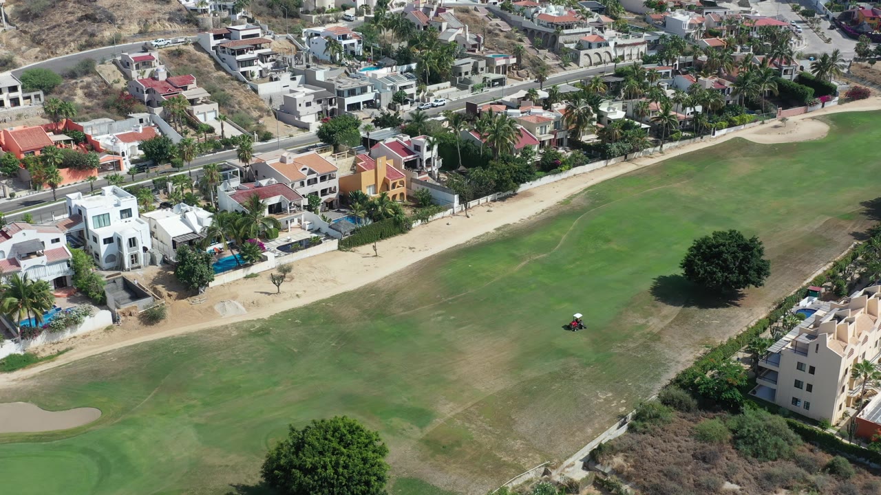 Aerial frame in Los Cabos, Mexico, with the tractor slightly right of center on the lawn, a large tree below, and rows of private homes along the upper edge.
