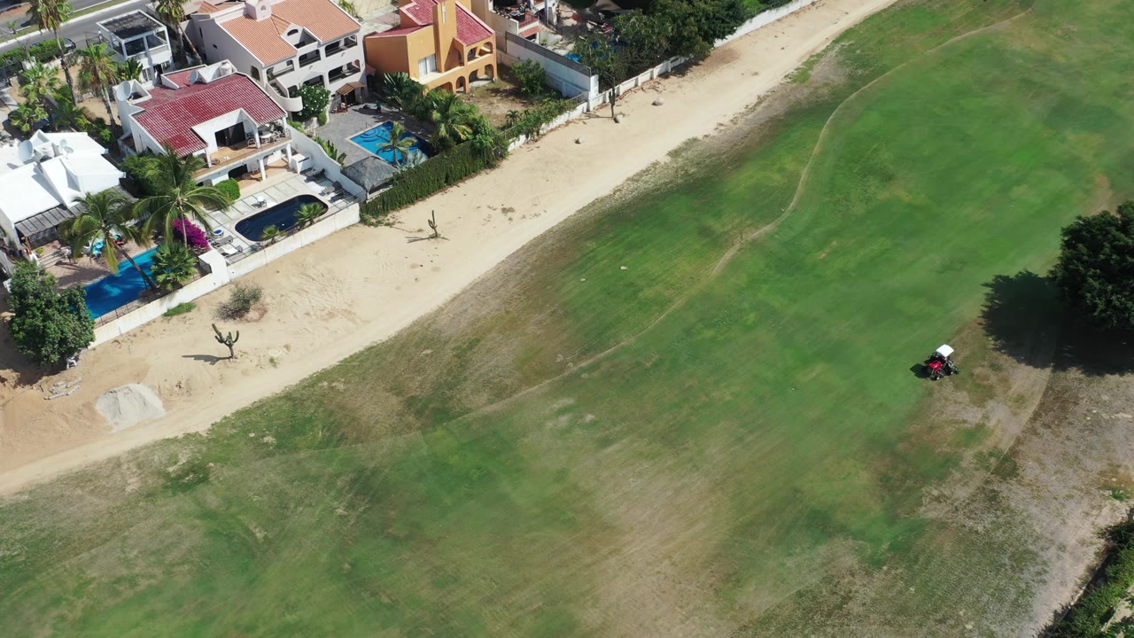 Aerial frame in Los Cabos, Mexico, with white and red-roofed homes at the upper left, a sandy path through the center, and green fairway-like ground to the right.