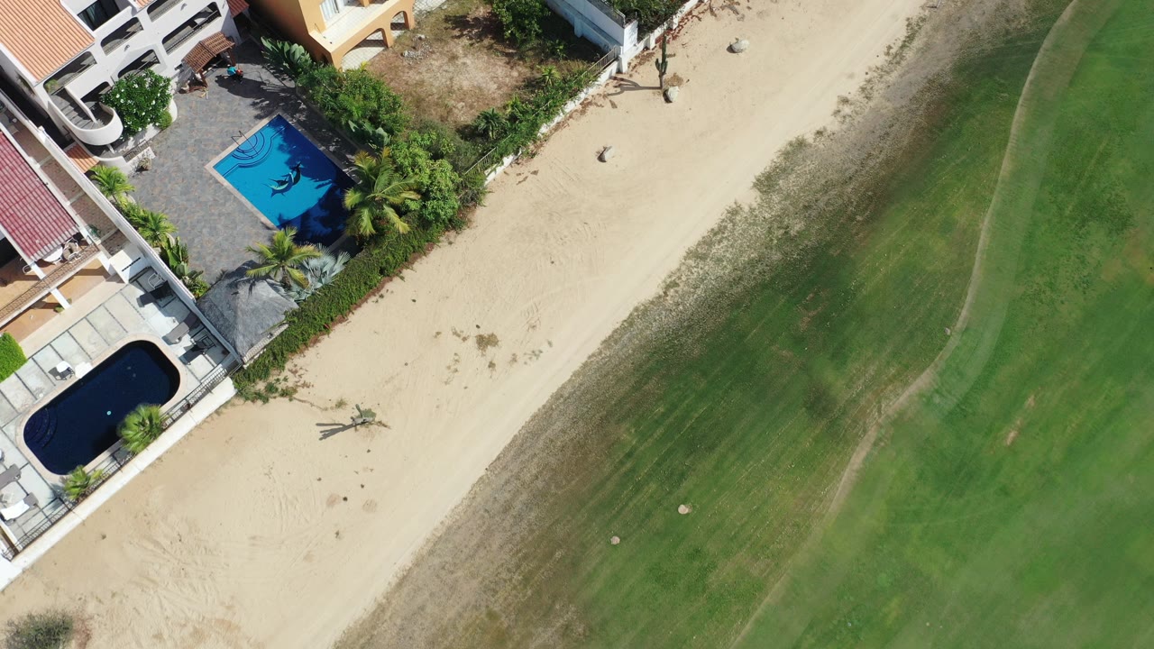 Overhead Los Cabos frame showing villas and two pools beside a tan beach and shallow green ocean, with no visible people.