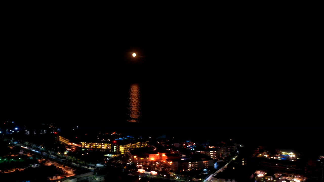 Full moon above the dark ocean in Los Cabos, Mexico, with a vertical orange reflection on the water and distant shoreline buildings lit below, no visible people.