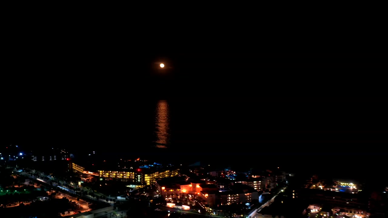The moon remains high over black water in Los Cabos, Mexico, with its reflection centered above a band of warm building lights and road lights, no visible people.