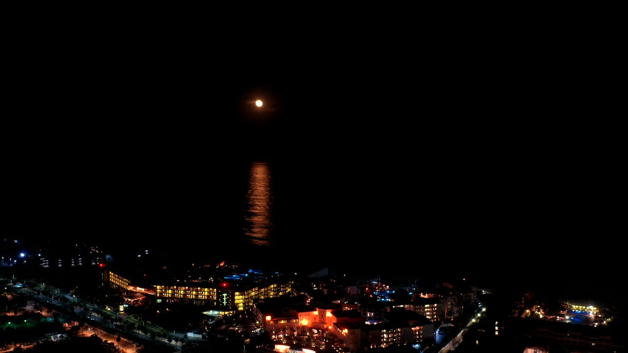 Night aerial view of moon over water and lit shoreline.