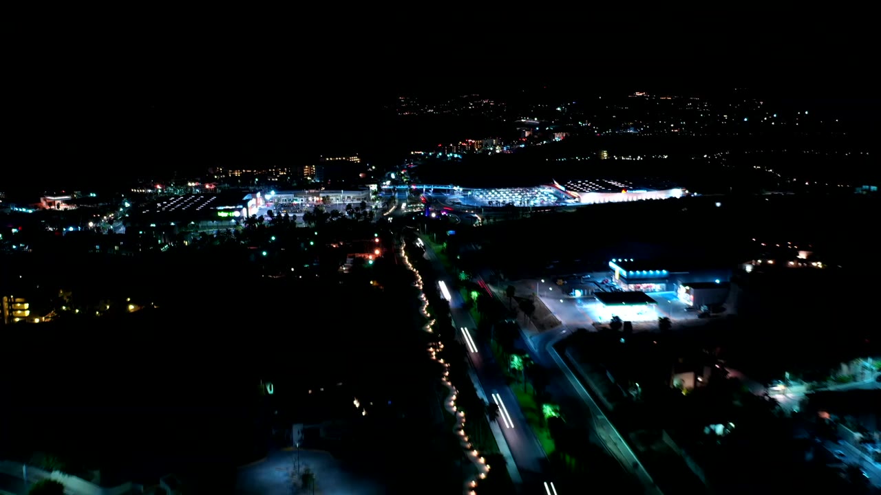 Los Cabos night aerial frame with a bright commercial-looking complex, curving roads, small light trails, dark surrounding land, and no visible people.