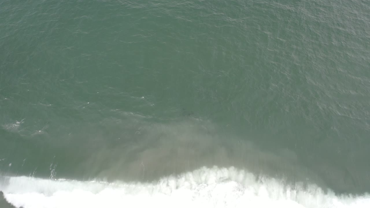 Blue-green water fills the overhead frame in Los Cabos, Mexico, while white foam spreads wider across the lower edge and no visible people appear.