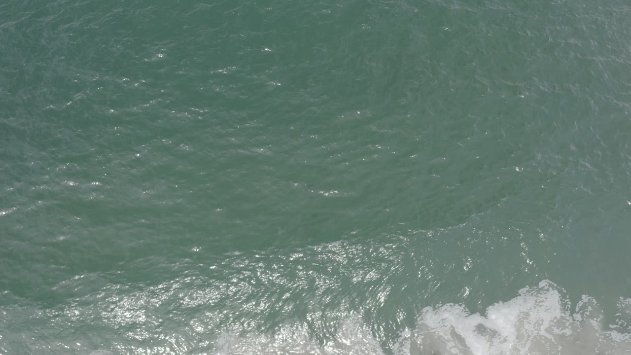 Top-down view of teal ocean water in Los Cabos, Mexico, with sunlight glinting on ripples, a white foam band near the bottom edge, and no visible people.